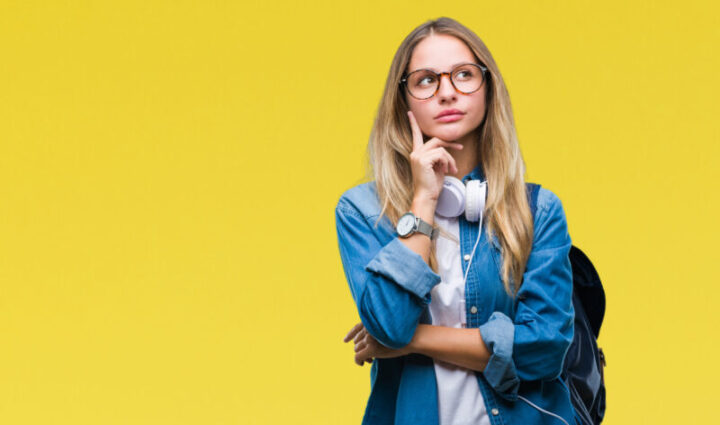 Young beautiful blonde student woman wearing headphones and glasses over isolated background with hand on chin thinking about question, pensive expression. Smiling with thoughtful face. Doubt concept.