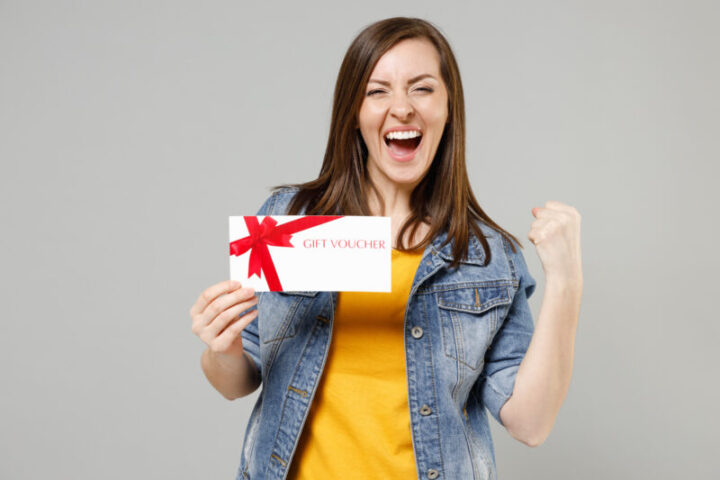 Young happy caucasian woman 20s in casual trendy denim jacket yellow t-shirt holding in hand gift voucher flyer mock up look camera clench fist do winner gesture isolated on grey background studio