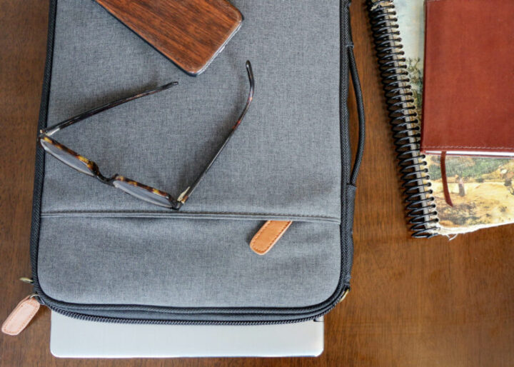 grey laptop case sits on brown table next to old book and notepad and journal ready for digital nomad to begin entrepreneur business. laptop case is soft and has zipper with pocket on laptop case.